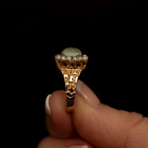 May include: A gold ring featuring a light green oval stone surrounded by small white pearls. The ring's band has an ornate, textured design. The ring is set against a black background.