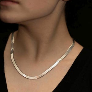 May include: A silver-toned herringbone chain necklace featuring a flat, flexible, and tightly woven link design that rests against the neck. The metallic surface reflects light, showcasing a smooth, continuous texture throughout the entire length of the chain.