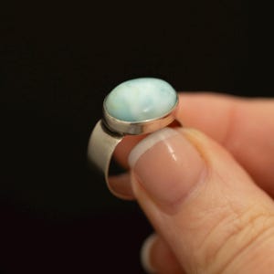 Könnte beinhalten: Ein Silberring mit einem glatten, ovalen, hellblauen Larimar-Edelstein, der in einer einfachen Metallfassung sitzt. Das Band des Rings ist breit und poliert und weist eine saubere, metallische Oberfläche auf, die den Stein ergänzt.
