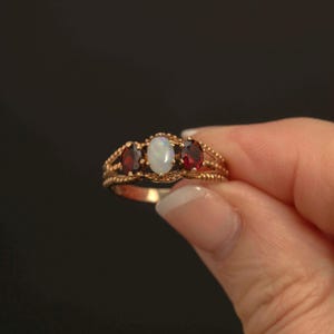 Könnte beinhalten: Ein goldener Ring mit drei ovalen Edelsteinen: ein Opal in der Mitte, flankiert von zwei Granaten. Der Ring hat ein detailliertes Filigran-Design. Die Edelsteine sind in einem goldenen Band gefasst.