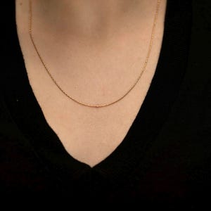 May include: A thin, delicate gold chain necklace with a fine link structure. The necklace rests against the neck, featuring a polished metallic finish that catches the light. The chain is simple, slender, and uniform in its design, creating a subtle look.