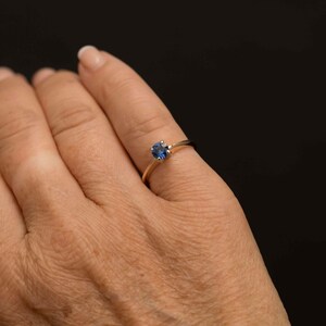 May include: A gold ring featuring a round, deep blue gemstone. The ring is set on a slender band and is shown on a person's finger against a black background. The gemstone appears to be approximately 0.6 cm in diameter.