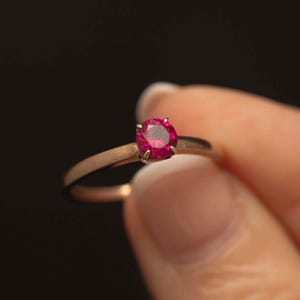 Könnte beinhalten: Ein goldener Ring mit einem runden, leuchtend roten Edelstein. Der Ring hat ein schmales Band, und der Edelstein ist der Mittelpunkt, gefasst in einer klassischen Vier-Krappen-Fassung. Der Ring wird vor einem dunklen Hintergrund gehalten.