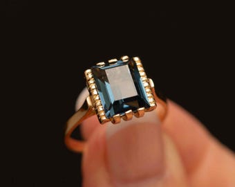 Può includere: Un anello d'oro con una grande pietra preziosa blu rettangolare. La pietra preziosa è incastonata in una montatura d'oro con un bordo decorativo. La fascia dell'anello è in oro lucido semplice. L'anello è mostrato su uno sfondo scuro.