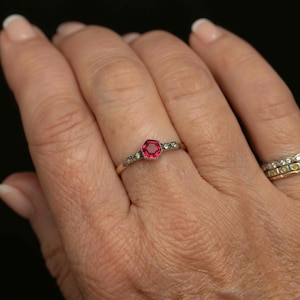 May include: A gold ring featuring a hexagonal, ruby-colored gemstone as the centerpiece. The band is adorned with small, clear gemstones. The ring is displayed on a hand, showcasing its design and details.