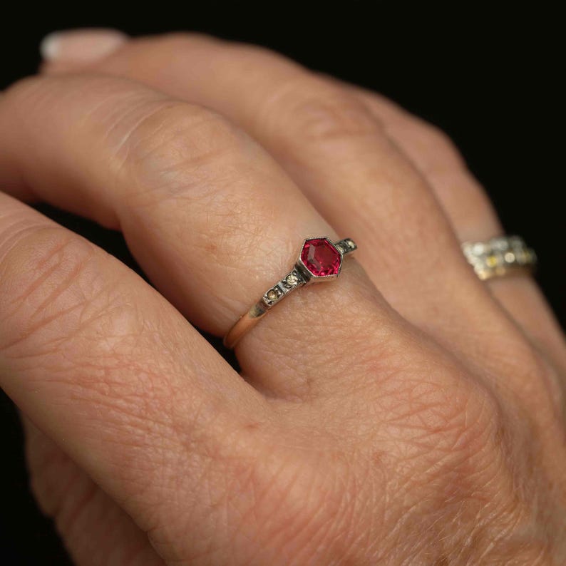 May include: A gold ring featuring a hexagonal, deep red gemstone as the centerpiece. The band is adorned with small, clear accent stones. The ring is set against a blurred background, highlighting the jewelry's details.