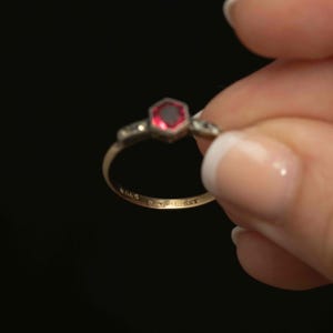 May include: A gold ring with a hexagonal, red gemstone at the center. The ring has small accent stones on either side of the central stone. The band is thin and has markings. The ring is held against a black background.