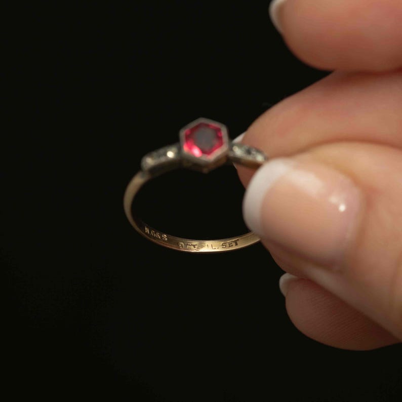 May include: A gold ring with a hexagonal, red gemstone at the center. The ring has small accent stones on either side of the central stone. The band is thin and has markings. The ring is held against a black background.