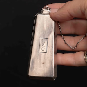May include: A silver-toned rectangular pendant with a chain. The pendant has a tapered shape with vertical lines and the letters "INC" in a rectangular box. The chain is delicate and silver. The background is black.