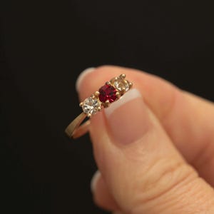 May include: A gold ring with three gemstones: a clear, a red, and a light-colored stone. The ring is held against a black background, showcasing the gemstones' sparkle. The ring's band is thin and delicate.