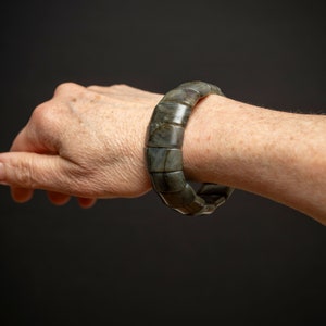 May include: A dark grey stone bracelet with a unique, segmented design. The bracelet is made of multiple pieces of stone that are connected together to form a continuous band.
