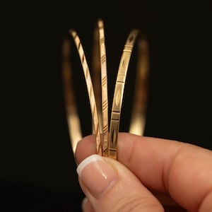 May include: Three thin gold bangles featuring distinct etched patterns. One bangle displays a diagonal line texture, another shows a cross-hatched design, and the third features a repeating pattern of horizontal lines and elongated diamond shapes.