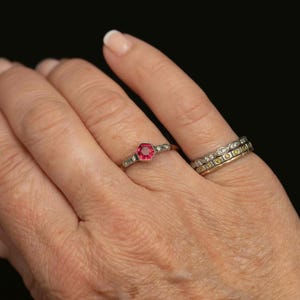 May include: A hand wearing two rings. The first ring has a hexagonal, bright red gemstone in the center, surrounded by small, clear stones. The second ring is a band with alternating clear and yellow stones.