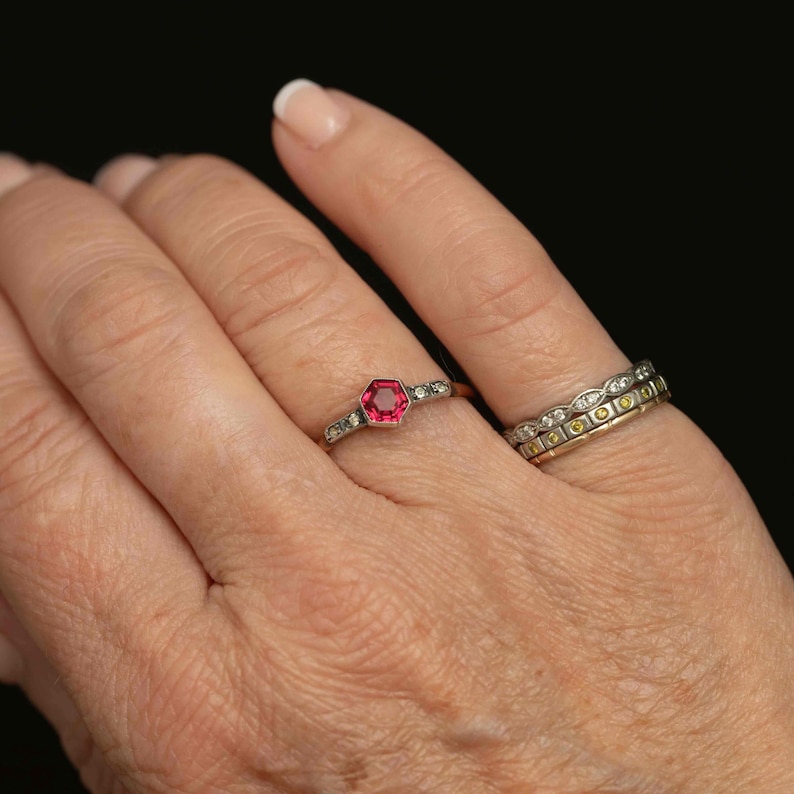 May include: A hand wearing two rings. The first ring has a hexagonal, bright red gemstone in the center, surrounded by small, clear stones. The second ring is a band with alternating clear and yellow stones.