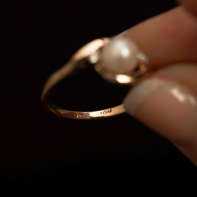 May include: A gold ring with a pearl and small accent stones. The ring has a delicate, curved design. The pearl is a creamy white color. The ring is held against a dark background.