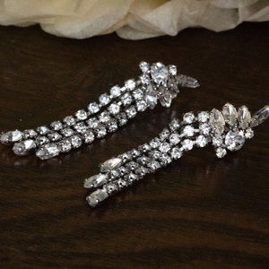May include: A pair of silver dangle earrings featuring clear crystal rhinestones. The earrings are made up of multiple strands of rhinestones that cascade down from a larger cluster of rhinestones at the top.