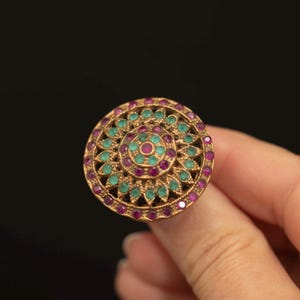May include: A circular gold-toned ring featuring a detailed floral-inspired design. The surface is adorned with numerous small, round, vibrant pink and green gemstones arranged in concentric patterns across the face of the jewellery piece.