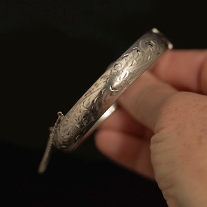 May include: A silver-toned hinged bangle bracelet featuring an intricate engraved floral pattern across its outer surface. The bracelet includes a secure clasp mechanism and a small attached safety chain for added security during wear.
