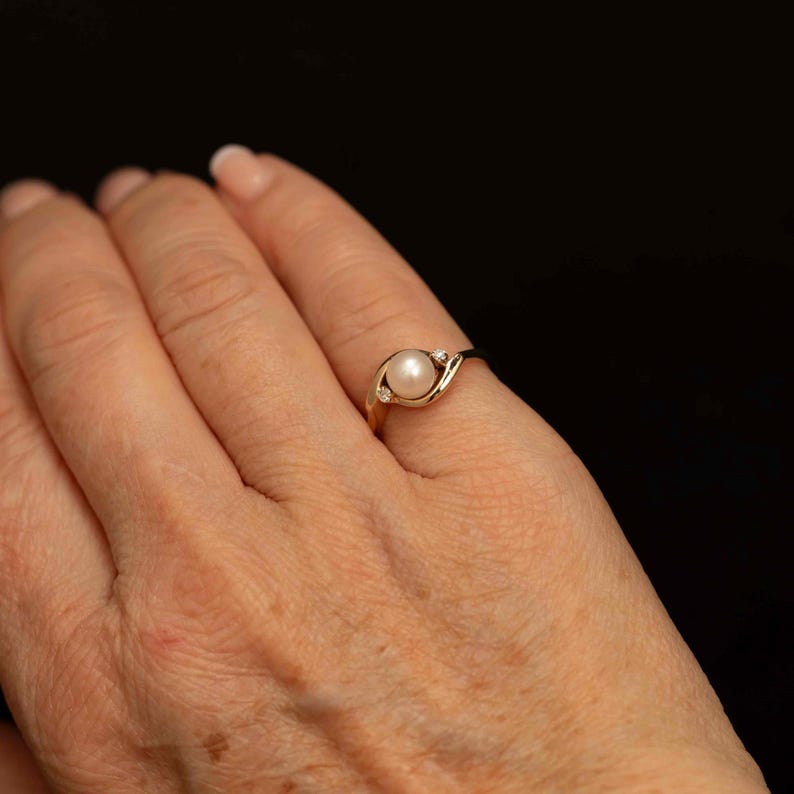 May include: A gold ring featuring a central pearl and a small diamond accent. The ring's band curves elegantly around the pearl, creating a delicate and classic design. The ring is shown on a person's finger.