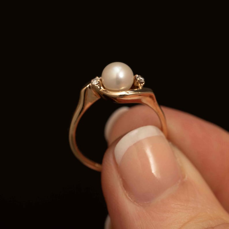 May include: A gold ring featuring a large, round, white pearl as the centerpiece. Two small accent diamonds flank the pearl. The ring has a delicate, curved design, and is held against a black background.