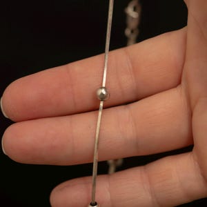 May include: A silver-toned snake chain necklace featuring small, evenly spaced round metal beads along its length. The chain has a smooth, flat, and flexible texture, with a metallic finish that reflects light, and a lobster claw clasp visible.