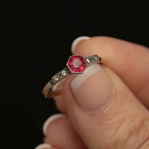 May include: A close-up of a vintage ring featuring a hexagonal, ruby-colored gemstone at the center. The ring has a silver band with small, clear accent stones on either side of the central stone. The ring is held against a dark background.