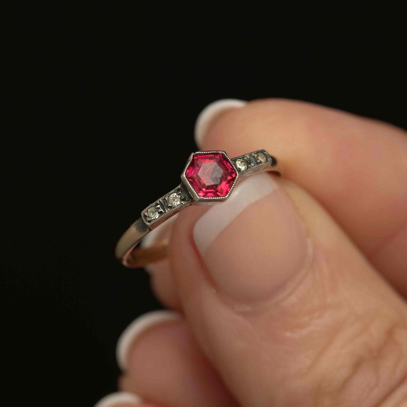 May include: A close-up of a vintage ring featuring a hexagonal, ruby-colored gemstone at the center. The ring has a silver band with small, clear accent stones on either side of the central stone. The ring is held against a dark background.