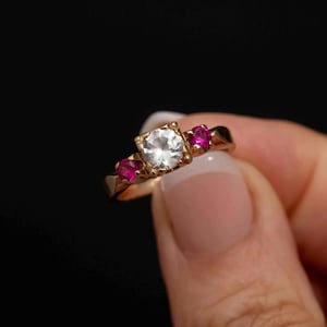 May include: A gold ring with a large, clear, round gemstone and two smaller, pink gemstones.