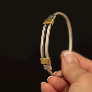 Könnte beinhalten: Ein kreisförmiger Armreif mit zwei parallelen silberfarbenen Bändern, die durch zwei rechteckige goldfarbene Metallakzente verbunden sind. Der Armreif hat eine glatte, polierte Oberfläche und eine starre Struktur in einem zweifarbigen Design.
