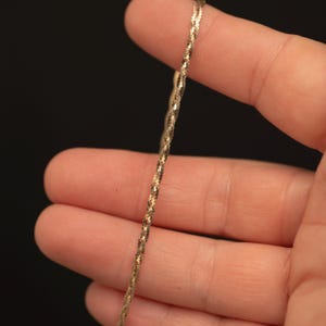 May include: A thin, gold-toned chain necklace featuring a twisted, textured link design. The metal surface has a reflective, shimmering quality that catches the light, creating a repeating pattern along the length of the delicate, slender jewellery piece.