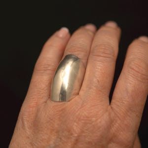 May include: A wide, polished silver-toned ring with a smooth, curved, and elongated surface that covers a significant portion of the finger. The metal has a reflective finish and a solid, continuous band design that tapers slightly towards the base.