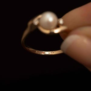 May include: A gold ring featuring a large, round, white pearl as the centerpiece. The ring has a delicate design with small accent stones on either side of the pearl. The ring is held against a black background.