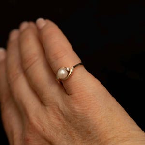 May include: A gold ring featuring a white pearl and two small diamonds. The ring has a delicate, swirling design. The ring is on a finger against a black background. The ring is a piece of jewelry.