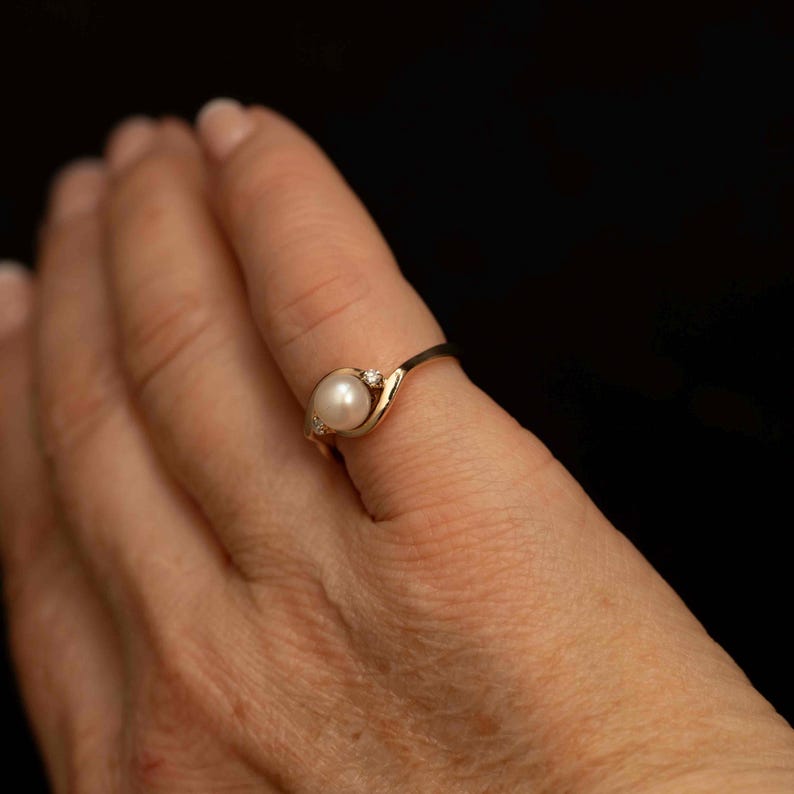 May include: A gold ring featuring a white pearl and two small diamonds. The ring has a delicate, swirling design. The ring is on a finger against a black background. The ring is a piece of jewelry.