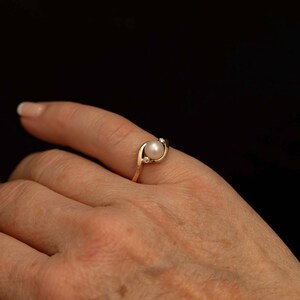 May include: A gold ring featuring a white pearl and two small accent diamonds. The ring has a delicate, swirling design, and is set against a dark background.