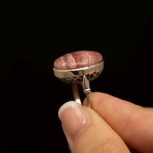 May include: A ring featuring a smooth, oval-shaped pink and white banded stone set in a silver-toned metal bezel. The setting displays a decorative, openwork honeycomb-like pattern along the side, supported by a simple, slender metal band.