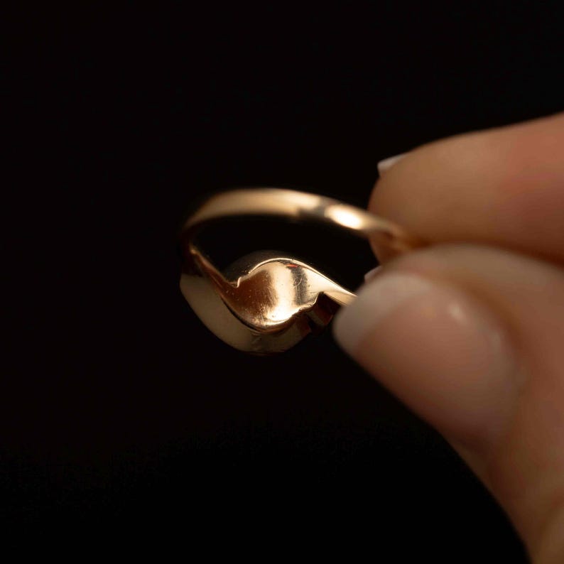 May include: A gold ring with a heart-shaped design. The ring is held against a black background, showcasing its polished surface and intricate curves. The ring's warm gold color contrasts with the dark backdrop, highlighting its elegant form.