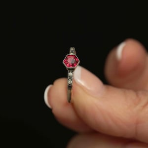 May include: A silver ring featuring a hexagonal, red gemstone. The band is detailed with small, clear accent stones. The ring is set against a black backdrop.