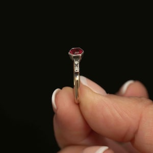 May include: A gold ring featuring a faceted, deep red gemstone as the primary focal point. The ring has a delicate band and is accented with two smaller, clear gemstones. The ring is held against a black background.