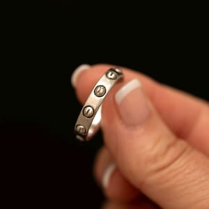 May include: A silver-toned metal ring featuring a series of evenly spaced, circular indentations along the outer band. Each circular detail contains a small, horizontal line, resembling the head of a flat-head screw, set against a smooth finish.