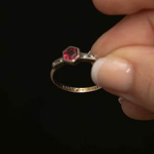 May include: A gold ring with a hexagonal red gemstone at the center, flanked by smaller clear stones. The ring has markings on the inside band, including "H.G.&S", "9CT", and "SET". The ring is held against a black background.