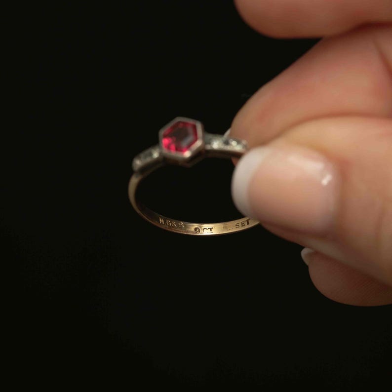 May include: A gold ring with a hexagonal red gemstone at the center, flanked by smaller clear stones. The ring has markings on the inside band, including "H.G.&S", "9CT", and "SET". The ring is held against a black background.