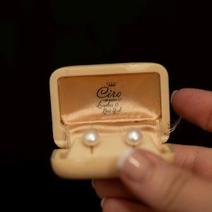 May include: A pair of white pearl stud earrings presented in a cream-colored rectangular jewelry box. The interior lid features the printed text Ciro of Bond St. London & New York with a small crown icon above the brand name on satin fabric.