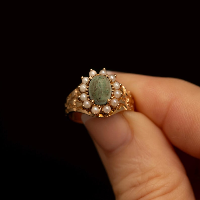 May include: A gold ring featuring an oval, green-hued stone surrounded by small, white pearls. The ring's band has an ornate, filigree design. The ring is held against a black background.