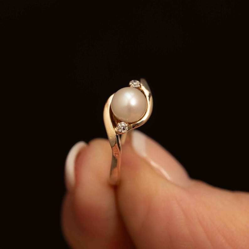 May include: A gold ring with a pearl center stone and two small accent diamonds. The ring has a swirling design, with the pearl set in the center and the diamonds on either side. The ring is held against a black background.
