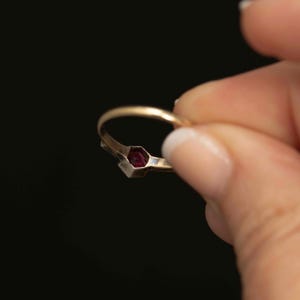 May include: A gold ring with a small, hexagonal, dark red gemstone. The ring is held against a black background, showcasing the ring's design and the gemstone's color.