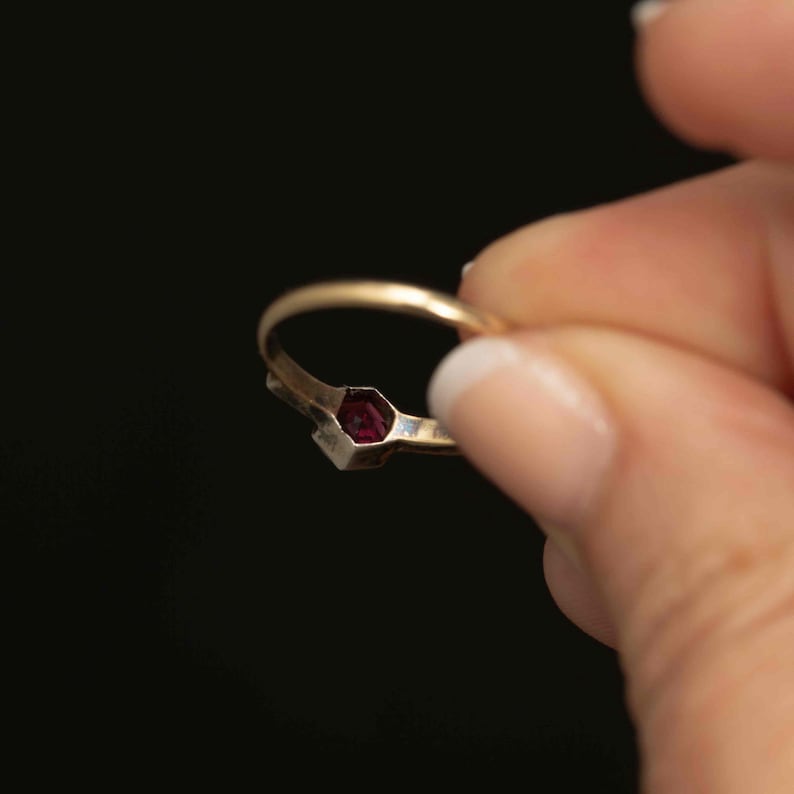 May include: A gold ring with a small, hexagonal, dark red gemstone. The ring is held against a black background, showcasing the ring's design and the gemstone's color.