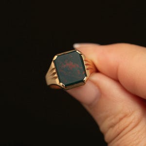 Könnte beinhalten: Ein goldener Ring mit einem rechteckigen, dunkelgrünen Blutstein mit deutlichen roten Einschlüssen. Das Metallband hat eine polierte Oberfläche und ein gestuftes, geometrisches Design an den Seiten, das zur zentralen Fassung führt.
