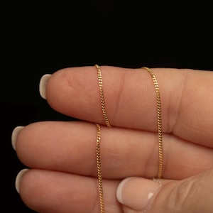 May include: A thin, delicate gold chain featuring small, interlocking links. The chain is draped across fingers, showcasing its reflective metallic surface and fine, uniform construction. The design consists of a continuous, slender metal strand.