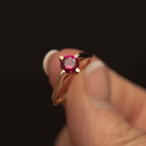 May include: A gold ring with a bright red gemstone. The ring has a simple, elegant design with a slender band and four prongs holding the gemstone. The ring is held against a dark background.